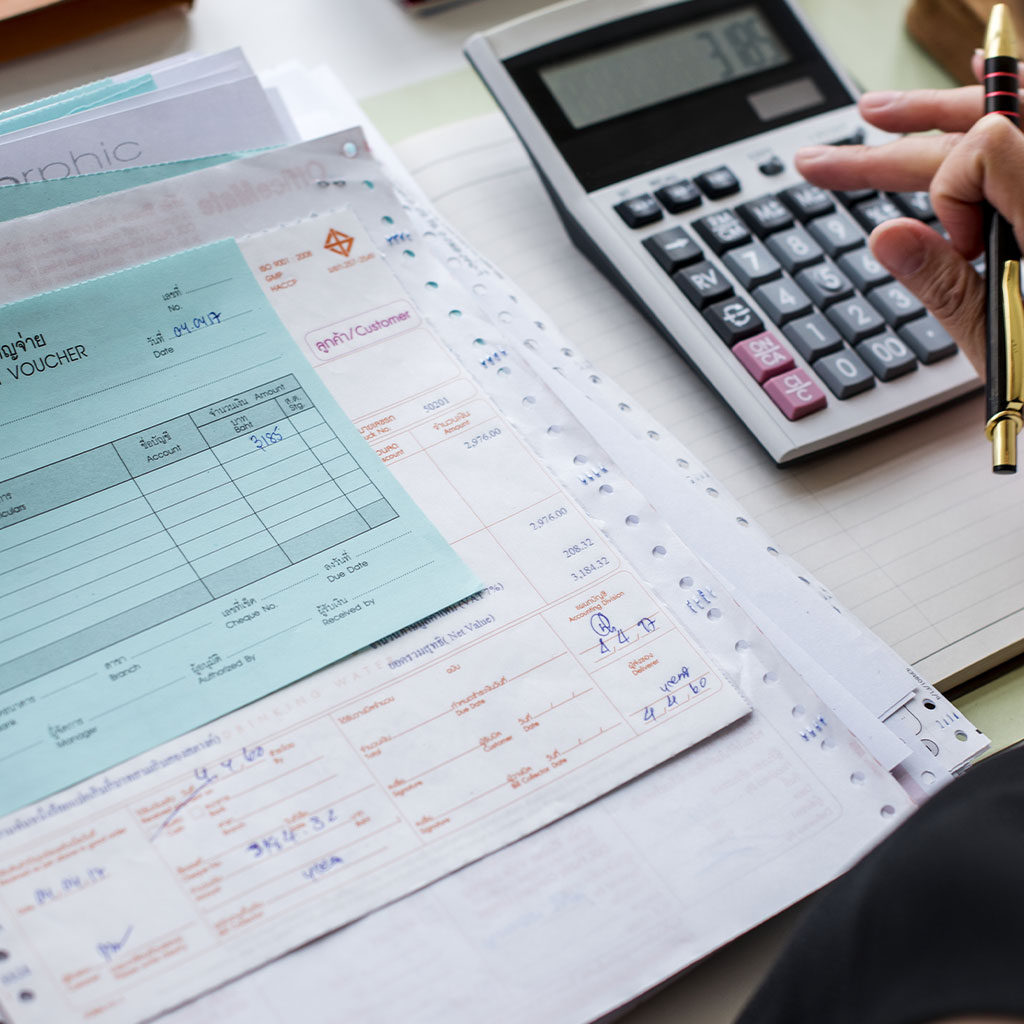 Una persona utilizando una calculadora con varias facturas, recibos y papeleo esparcidos sobre un escritorio, sosteniendo un bolígrafo y escribiendo en un cuaderno: captura perfectamente el trabajo orientado al detalle de la asesoría en Sevilla.