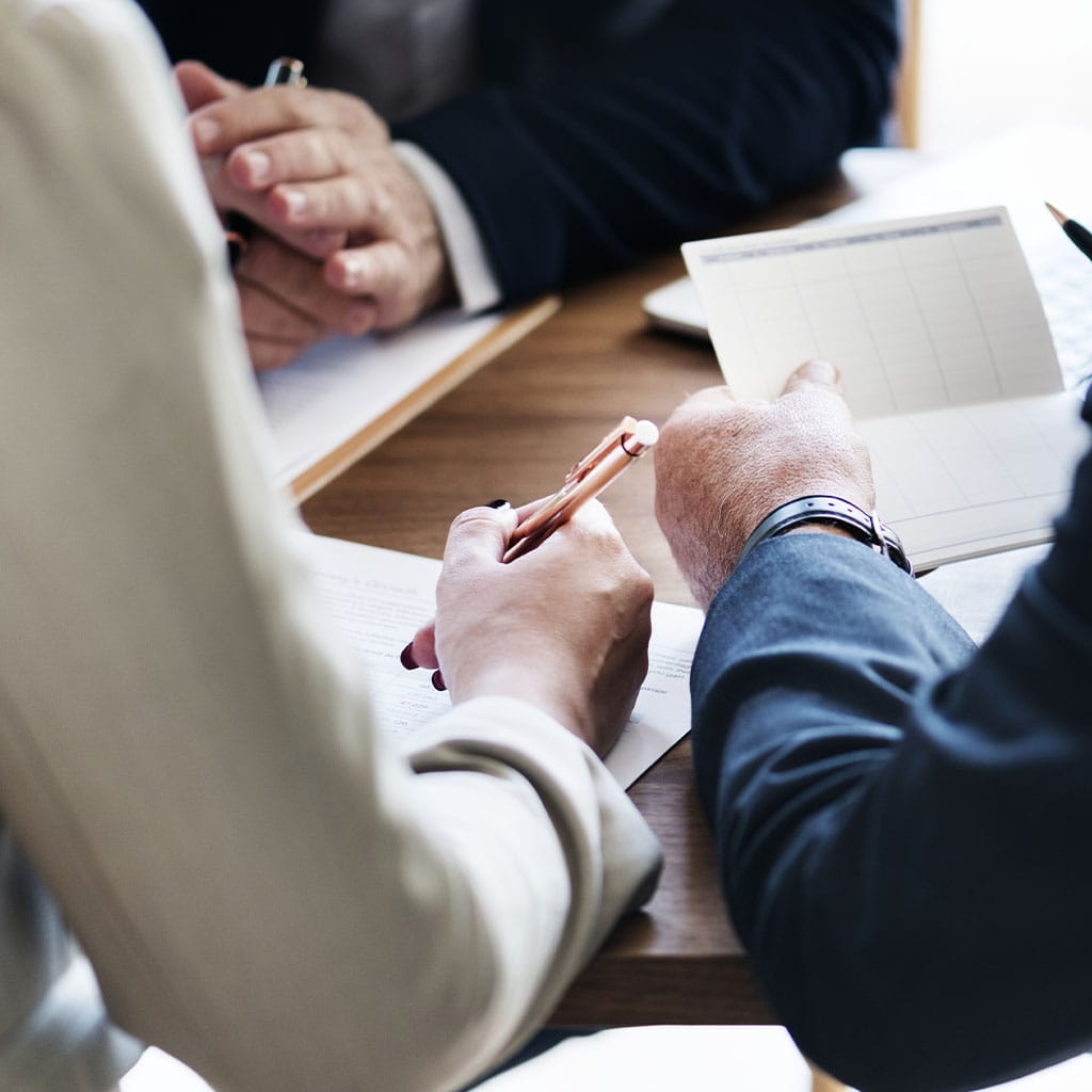 Tres personas en traje de negocios están sentadas en una mesa, discutiendo documentos. Una persona está escribiendo, otra sostiene un bloc de notas y una tercera tiene las manos entrelazadas durante una reunión de Asesoría en Sevilla.